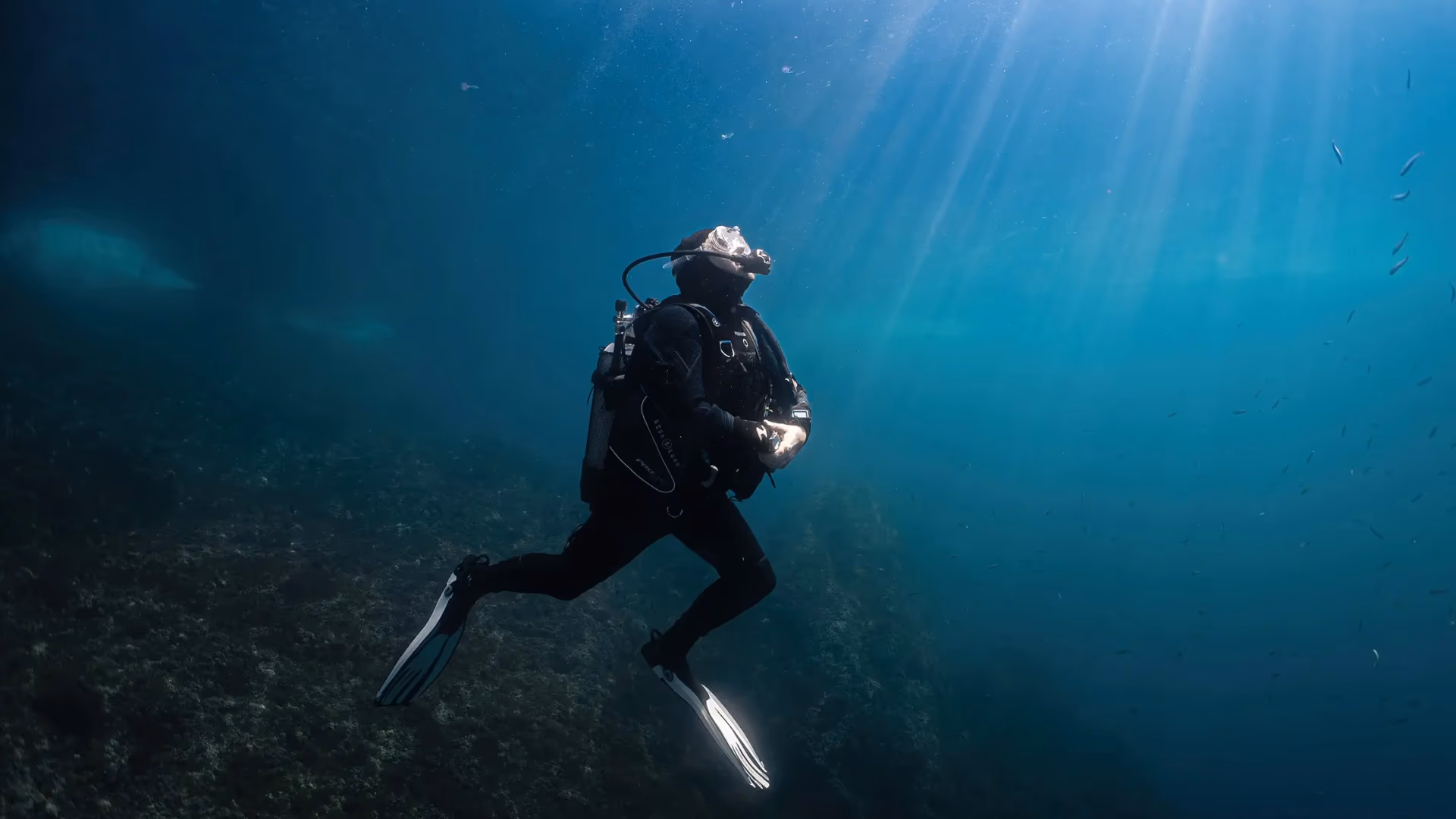 Diver in a diving suit moving underwater with sun rays filtering through the surface