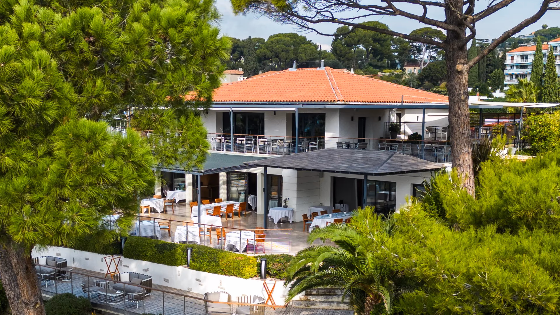 Restaurant with outdoor terrace and tables, surrounded by trees and vegetation, under a partly cloudy sky.