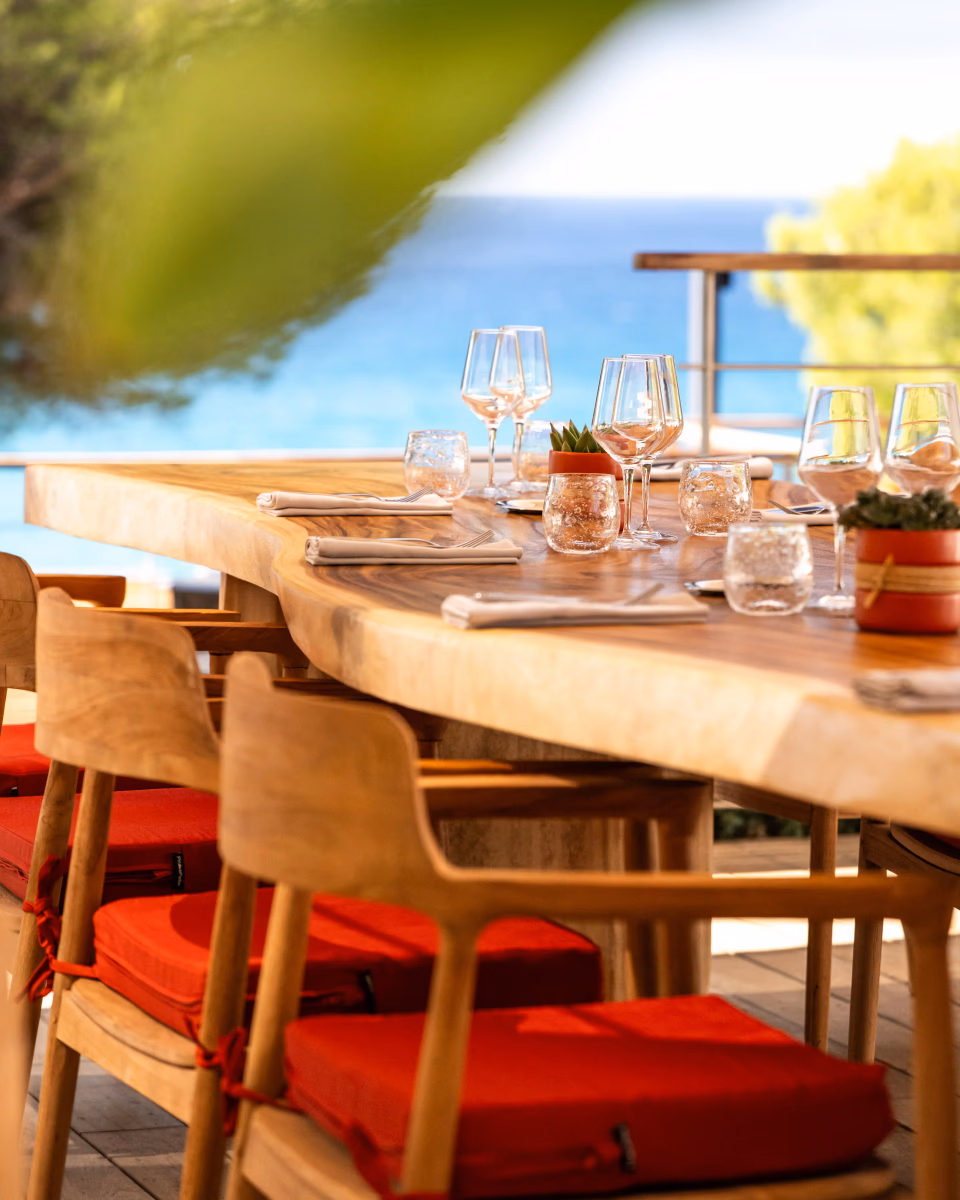 Wooden table with glasses, cutlery, and napkins, surrounded by wooden chairs with red cushions, outdoors near a body of water.