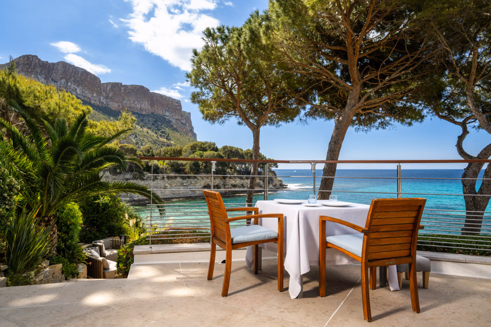 Table set with two wooden chairs on a terrace facing the sea, surrounded by trees and mountains.