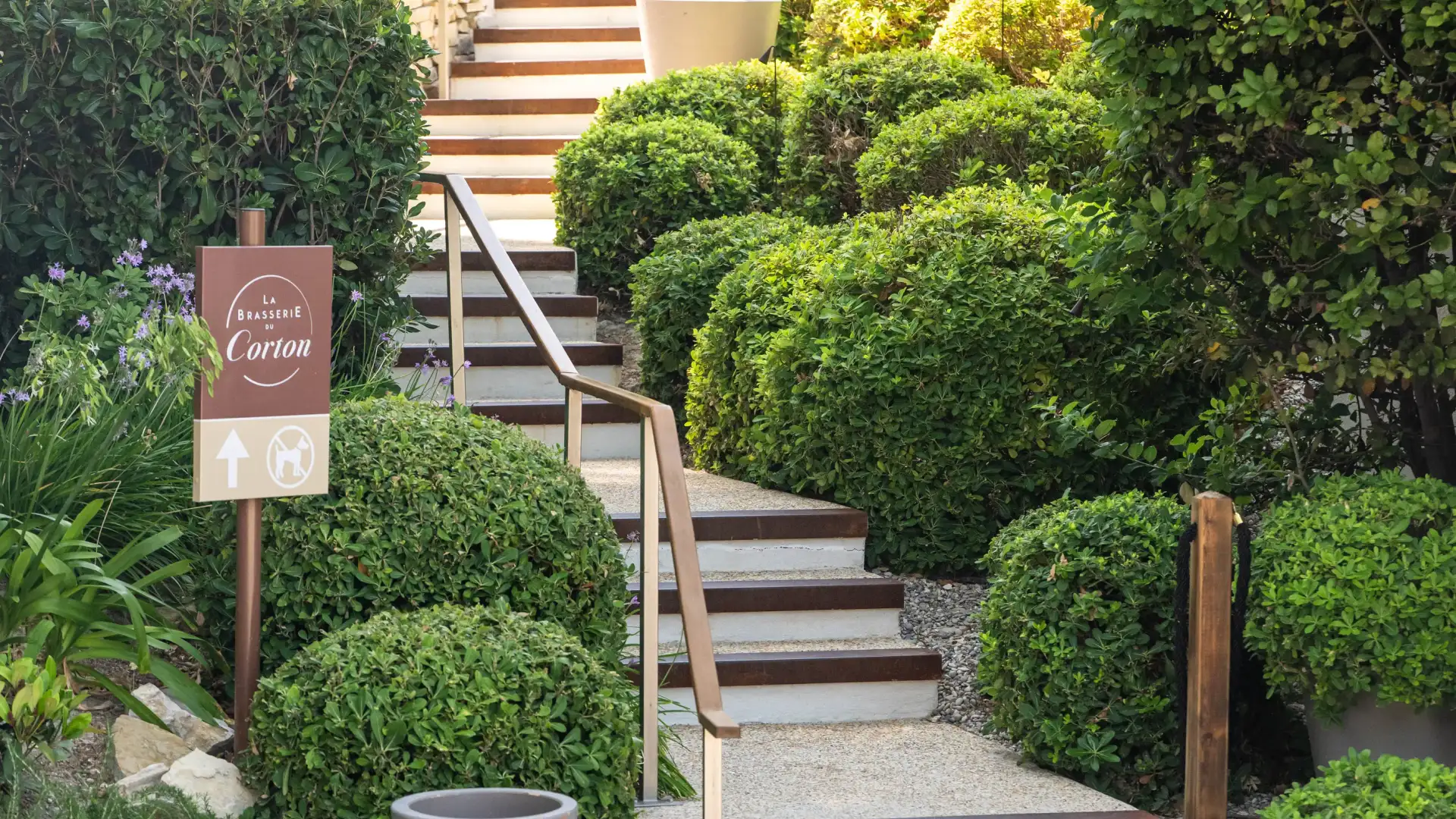 Un escalier en pierre avec des rampes en bois mène à une terrasse arborée, accompagné d'un panneau indiquant "La Brasserie Corom"