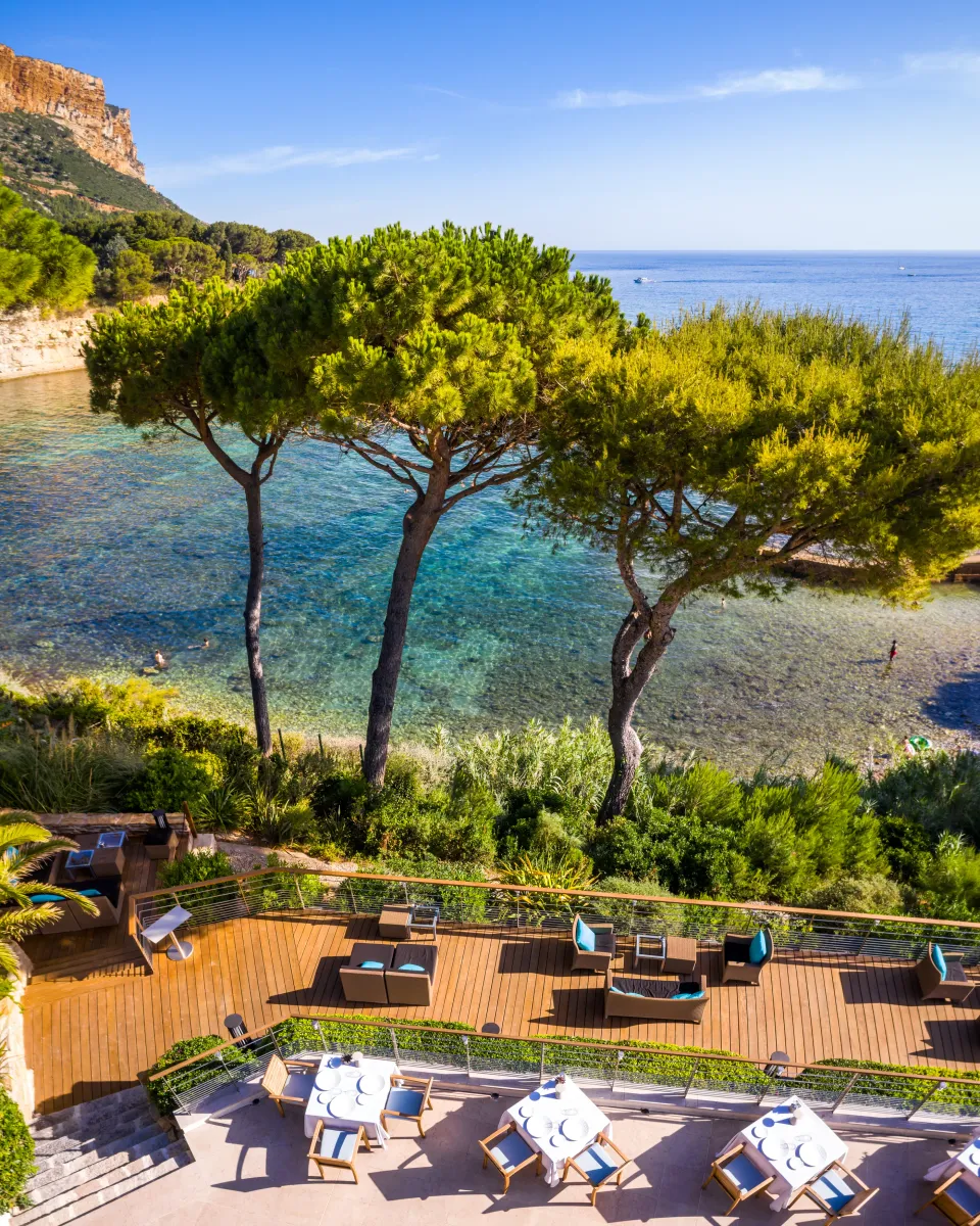 Terrasse en bois avec tables et chaises face à une crique bordée de pins et de végétation, avec une vue sur la mer bleue